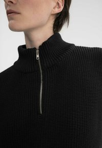 Schwarzer, gestrickter Pullover mit hohem Kragen, einer halben Reißverschlussöffnung und strukturiertem Stoff, der das gerippte Detail im gesamten Design betont.
