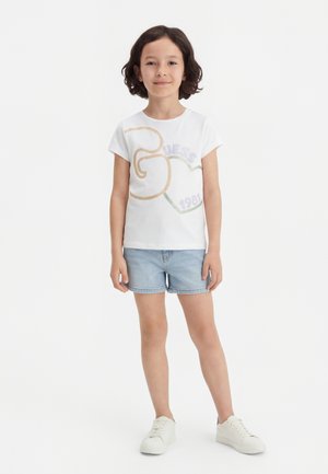 Fiatal gyermek rövid sötét hajjal, fehér mintás pólót, világos farmer rövidnadrágot és fehér tornacipőt viselve, egy sima fehér háttér előtt áll.