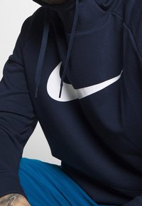 Granatowa bluza z kapturem Nike z dużym białym logo, cechująca się dopasowanym krojem, sznurkami do regulacji i fakturowaną tkaniną. W zestawie niebieskie spodnie.