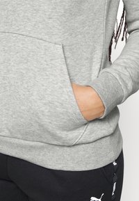 Personne portant un sweat-shirt gris clair avec la main dans la poche avant et un pantalon Puma noir avec logo visible sur la hanche.