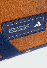 Borsa di tessuto arancione con superficie testurizzata, dotata di una tracolla blu e di un'etichetta rettangolare con testo e un'icona bianca.