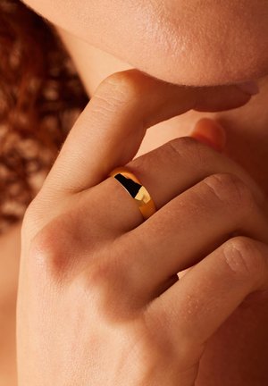 Gouden ring met een gladde, gepolijste oppervlakte, gedragen om een vinger. De band is eenvoudig en heeft een lichte glans, wat de huidskleur aanvult.