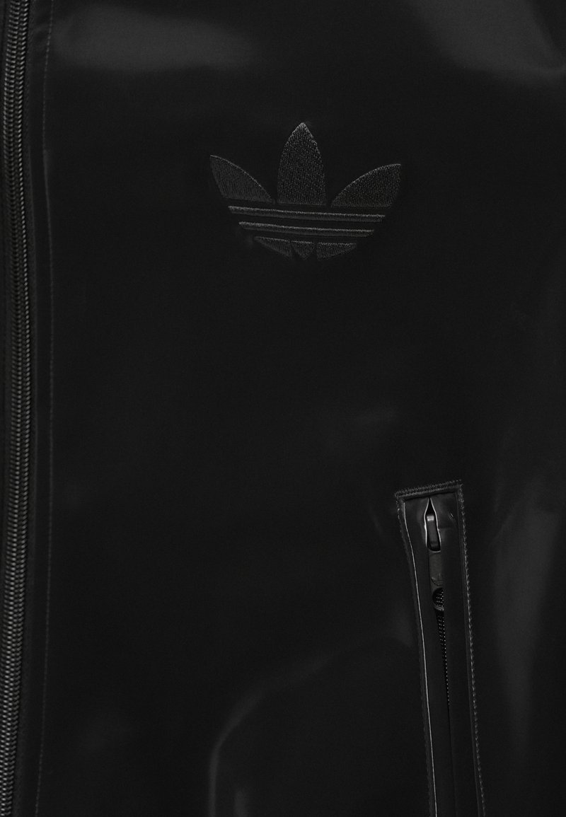 Schwarze Jacke mit Reißverschluss und einer vertikalen Reißverschlusstasche, mit einem gestickten Adidas Trefoil-Logo auf der Brust.