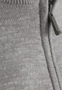 Grijze zip-up trui met een gestructureerd breipatroon. Kenmerkt een metalen ritsstopper met het label "SUITABLE." Close-up kijk benadrukt de stof en de steken.