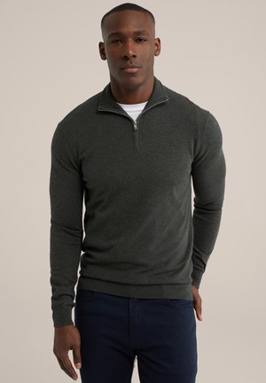 Dark green zip-up sweater in a soft knit material, featuring a quarter-length zipper and ribbed cuffs. Worn with navy pants.