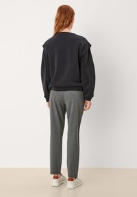 Persona con cabello rojo vistiendo un sudadera negra de gran tamaño, pantalones rectos grises y zapatillas blancas con detalles en marrón, de pie contra una pared sencilla.