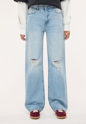 Jeans azzurri chiari a gamba larga con ginocchia strappate indossati con sneakers bordeaux e top a manica lunga neri e bianchi a strati.