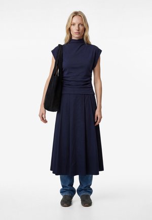 Blonde Frau trägt ein marineblaues ärmelloses Oberteil und einen Rock über blauen Jeans, schwarze Turnschuhe, trägt eine schwarze Umhängetasche vor weißem Hintergrund.