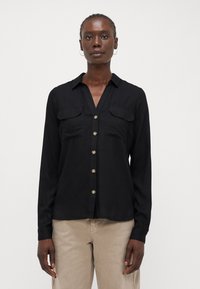 Chemise noire à manches longues avec col, deux poches poitrine et boutons dorés, fabriquée dans un tissu lisse et léger.
