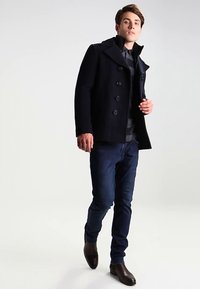 Marineblaue Peacoat mit strukturierter Stoffoberfläche und großen Knöpfen, kombiniert mit dunklen Jeans und braunen Lederstiefeln, mit einer maßgeschneiderten Passform und hohem Kragen.