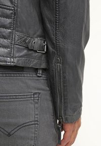 Sort læderjakke med quiltet skulderdetalje, lynlåse ved ærmerne og en justerbar rem ved taljen. Båret over grå denim.