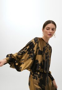 Zwarte en gouden tie-dye blouse met ballonmouwen, gemaakt van een gladde, glanzende stof. Heeft een geplooide manchet en een relaxte pasvorm.