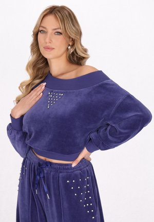 Femeie cu păr lung blond, purtând un hanorac croptat din velur violet și pantaloni asortați cu detalii din cristale strălucitoare.
