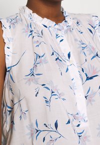 Blusa bianca con stampa floreale blu e rosa, colletto arricciato e chiusura frontale con bottoni. Tessuto leggero con pieghe vicino alle spalle.
