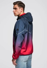 Mladý muž ve slunečnicích přechodových barev od modré po červenou s kapucí, v světlých modrých džínech, čelí vlevo s neutrálním výrazem.