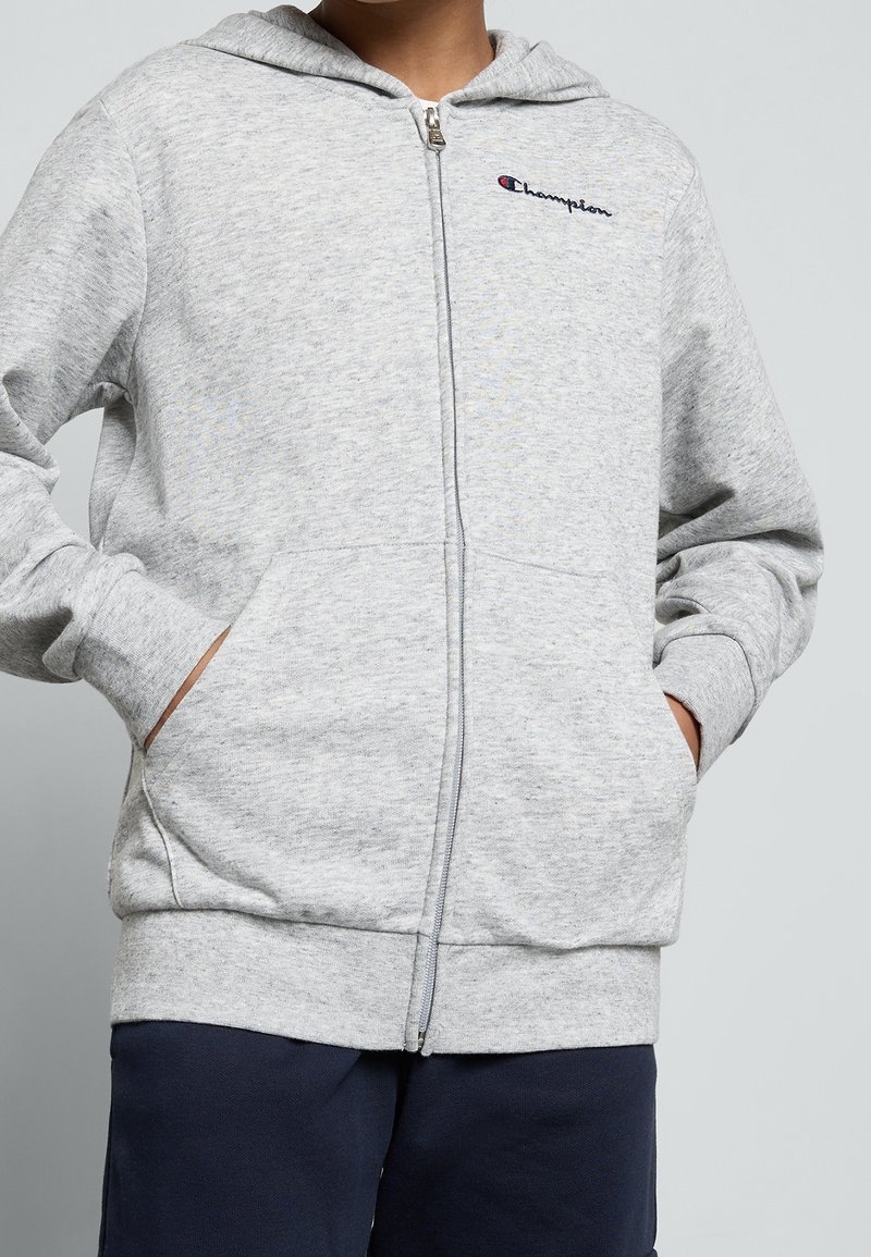 Sweat à capuche gris à fermeture éclair, fabriqué en tissu doux et texturé. Comprend une fermeture éclair devant, des poches kangourou, et un petit logo brodé sur la poitrine.