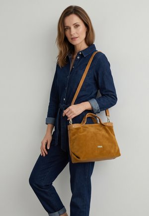 Frau in blauem Denim-Overall, die eine hellbraune Ledertasche mit Schulterriemen vor einem schlichten hellen Hintergrund trägt.