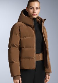 Smeđa puffer jakna s velikim kapuljačom, quiltanom teksturom i zatvaračem. Uparena s uskim crnim pletenim topom i crnim hlačama visokog struka.