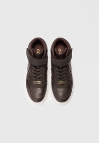Een paar donkerbruine GUESS high-top sneakers met witte zolen, veters en gemerkte bandjes op een plain witte achtergrond.