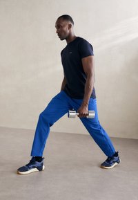 Sportos összeállítás, amely sötétkék pólót és kék nadrágot tartalmaz. Egy személy ezüst súlyt emel edzésállásban. Texturált sportcipők.