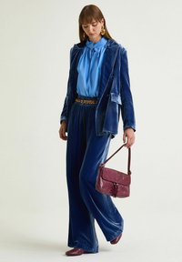 Abito blu vellutato con pantaloni a gamba larga e blazer abbinato. Combinato con una camicetta azzurro chiaro, cintura dorata, borsa bordeaux e accessori.