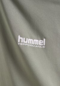 Gros plan sur un tissu vert texturé avec le logo blanc "hummel PERFORMANCE" imprimé sur la surface.