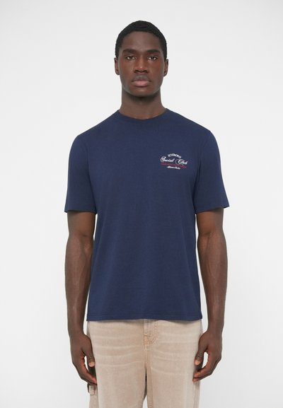 T-shirt en coton bleu marine à manches courtes, col rond, avec un petit logo brodé sur la poitrine, porté avec un jean de couleur claire.