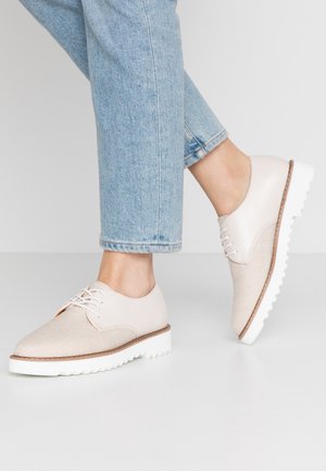 Cream-colored lace-up shoes with a textured fabric toe and smooth leather sides, featuring a white chunky sole and brown accents along the edge.