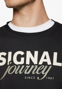 Sort sweatshirt med ribbet rund hals, der har stor hvid og lys grå tekst, der står "SIGNAL rejse Siden 1967."