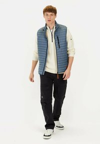 Blau gesteppte Weste über einem weißen Pullover, kombiniert mit schwarzen Cargo-Hosen und weißen Sneakers. Verfügt über Stickdetails und eine Reißverschlusstasche.