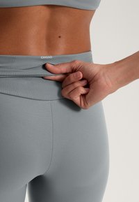 Sivé legíny s vysokým pásom, hladkou textúrou a jemnými švovými detailmi. Ruka uchopila látku pri páse, čo ukazuje na flexibilitu.