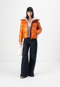 Puffer jacket in shiny orange material, with a furry collar and burgundy zipper, paired with wide-leg dark denim pants and a small white bag.