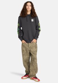 Jongere staat met handen in de zakken, draagt een zwarte hoodie met groene bladringen op de mouwen, gemusterde bruine broek en rode sneakers.