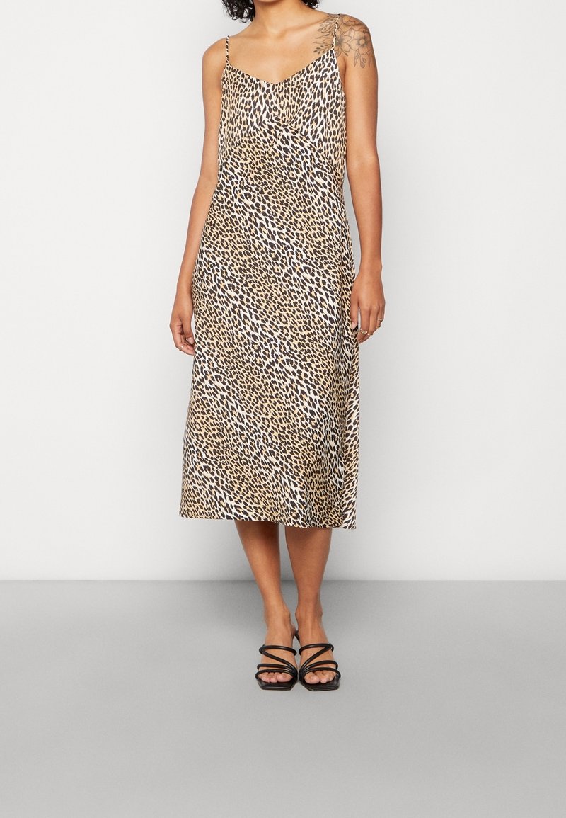 Femme portant une robe midi sans manches à imprimé léopard avec fines bretelles et sandales noires à brides, debout devant un fond uni.