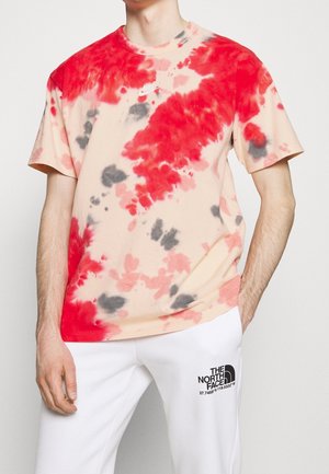 Mann trägt ein rot, pink und grau batik Nike T-Shirt und weiße Hose mit The North Face Logo am linken Oberschenkel.