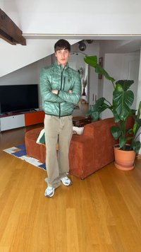Νέος άνδρας με πράσινο μπουφάν και μπεζ παντελόνι στέκεται με σταυρωμένα τα χέρια σε μοντέρνο σαλόνι με πορτοκαλί καναπέ και μεγάλο πράσινο φυτό.