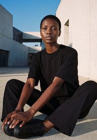 Young Black person with short hair wearing black clothes and shoes, sitting on the ground with legs apart against modern concrete buildings.