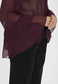 Burgundi chiffon blúz fodros hosszú ujjakkal, réteges textúrával és áttetsző kivitelben, fekete struktúrált nadrággal párosítva.