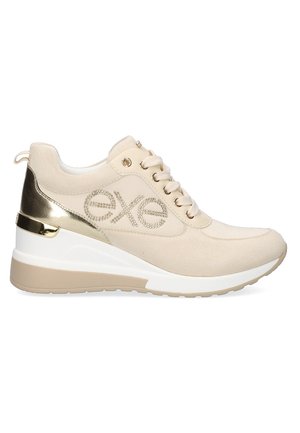 Zapatilla deportiva beige con textura de ante, detalles en dorado metálico, cierre con cordones y tacón en cuña; presenta detalles decorativos del logotipo.