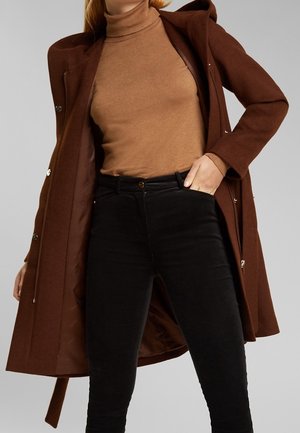 Kobieta w brązowym golfie i ciemnobrązowym otwartym płaszczu, nosząca czarne spodnie z wysokim stanem, z ręką w kieszeni na jednolitym tle.