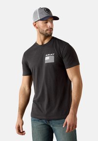 Camiseta negra de algodón con mangas cortas, con un logo blanco y un gráfico de la bandera americana en el pecho izquierdo. Se lleva con vaqueros azules y una gorra gris.