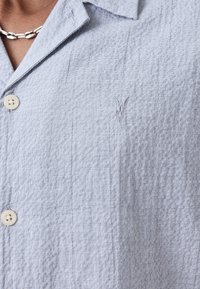 Camicia button-up azzurro chiaro realizzata in tessuto testurizzato, dotata di colletto, bottoni frontali e un piccolo logo ricamato sul petto.