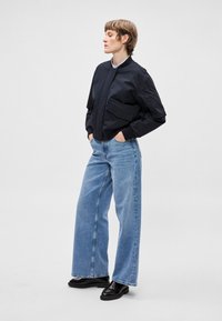 Tumesinine pommitaja jakk esiosa lukuga, suure tasku ja elastsete mansettidega, kombineeritud hele siniste kõrge vöökohaga laiade pükstega ja mustade loafers-kingadega.