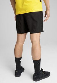 Sorte shorts med et enkelt design, parret med sorte ribbede sokker og sorte atletiske sko. Outfittet inkluderer en lys gul skjorte.