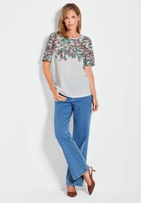 Lichtgrijze blouse met kleurrijke bloemenborduursels op de schouders, gecombineerd met wijde blauwe denim jeans en bruine hakken. Gladde textuur.