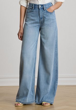 Persona che indossa jeans azzurri a gamba larga con vita alta, camicia bianca con maniche arrotolate e sandali gialli aperti in punta.