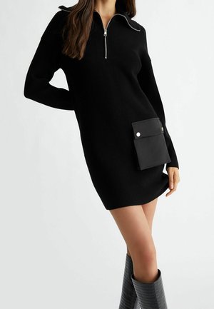 Donna che indossa un vestito nero a coste in maglia con collo a mezza cerniera, grande tasca laterale con pattine con bottone e stivali testurizzati al ginocchio.