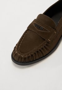 Brune suède loafers med samlet detaljering, en svart logobadge og kontrastsøm. Rund tå og svart gummisåle.