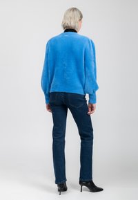 Person mit kurzen blonden Haaren, die einen leuchtend blauen, flauschigen Pullover, dunkle blaue Jeans und schwarze, hohe Stiefel trägt, steht mit dem Rücken zu einem schlichten Hintergrund.
