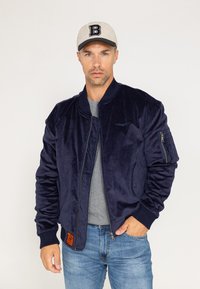 Veste bomber bleu marine en matériau doux, dotée d'une fermeture éclair à l'avant, de poignets côtelés et d'un détail en patch à la taille. Portée avec un t-shirt gris et un jean bleu.